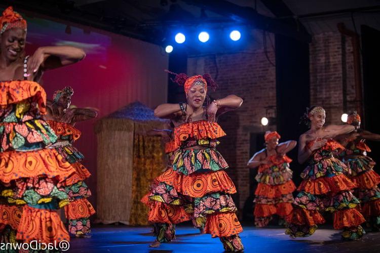 Photo of five dancers in bright orange African print costumes. The troupe is on a stage with three blue stage lights abover their heads. 