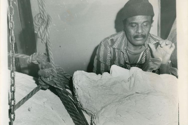 Black and white photograph of a Black man wearing a hat working on a sculpture of Frederick Douglass